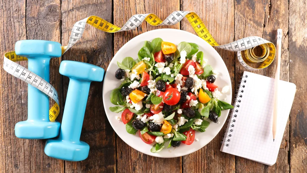 Fresh salad, dumbbells and a notebook on a wooden table